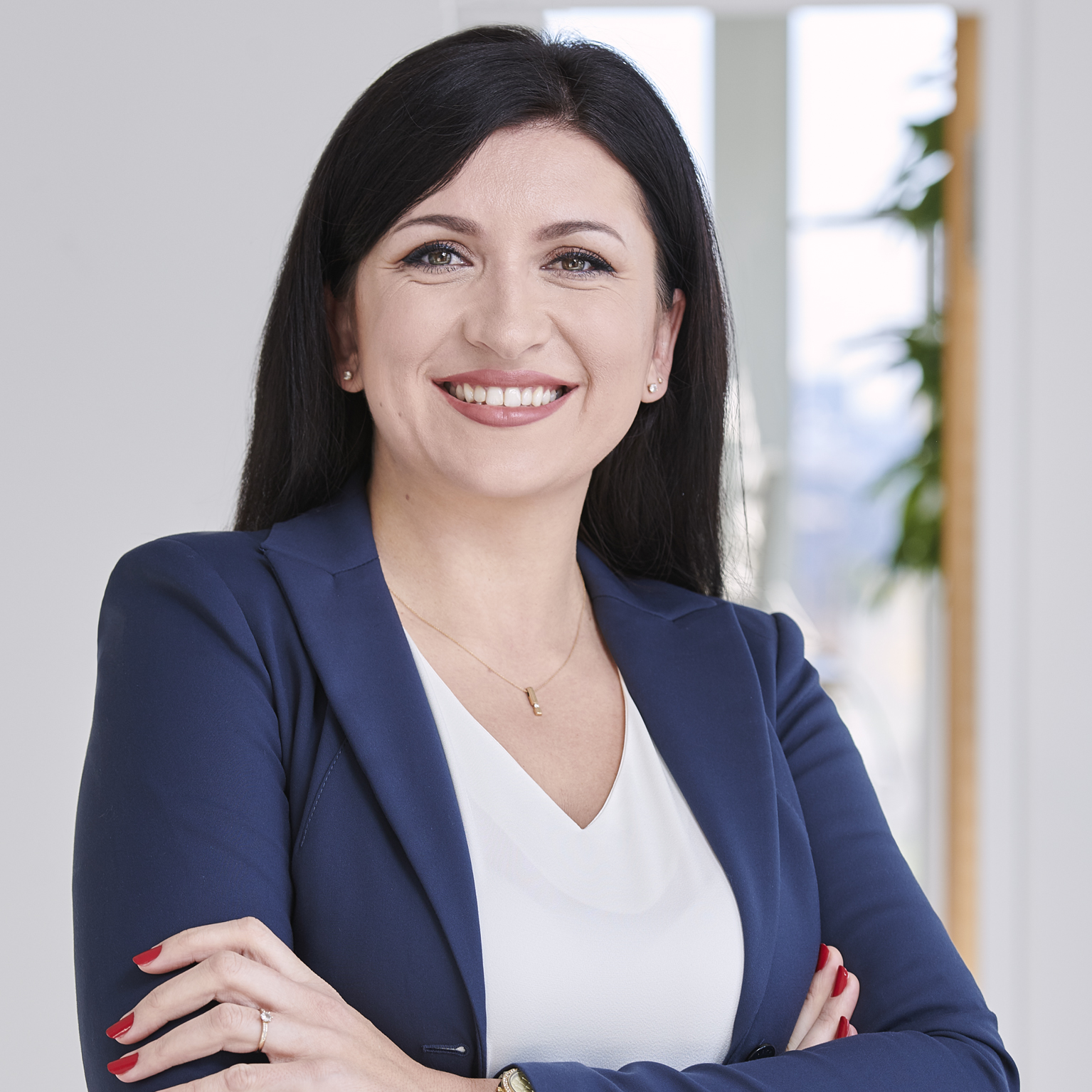 Portrait von Armina Kafedzic-Briga, lächelnd, in einem Business-Outfit vor einem neutralen Hintergrund mit Pflanzen.